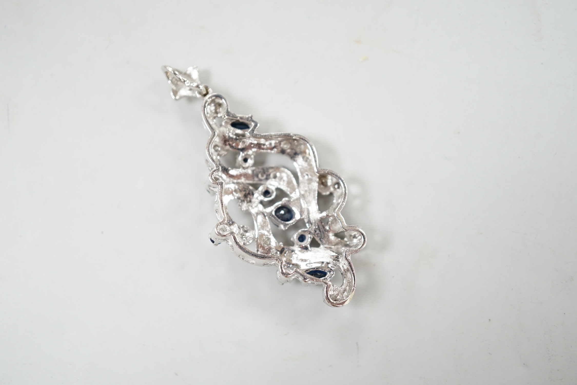 A modern 18k white metal sapphire and diamond chip cluster set scroll pendant, overall 47mm, gross weight 7.7 grams.